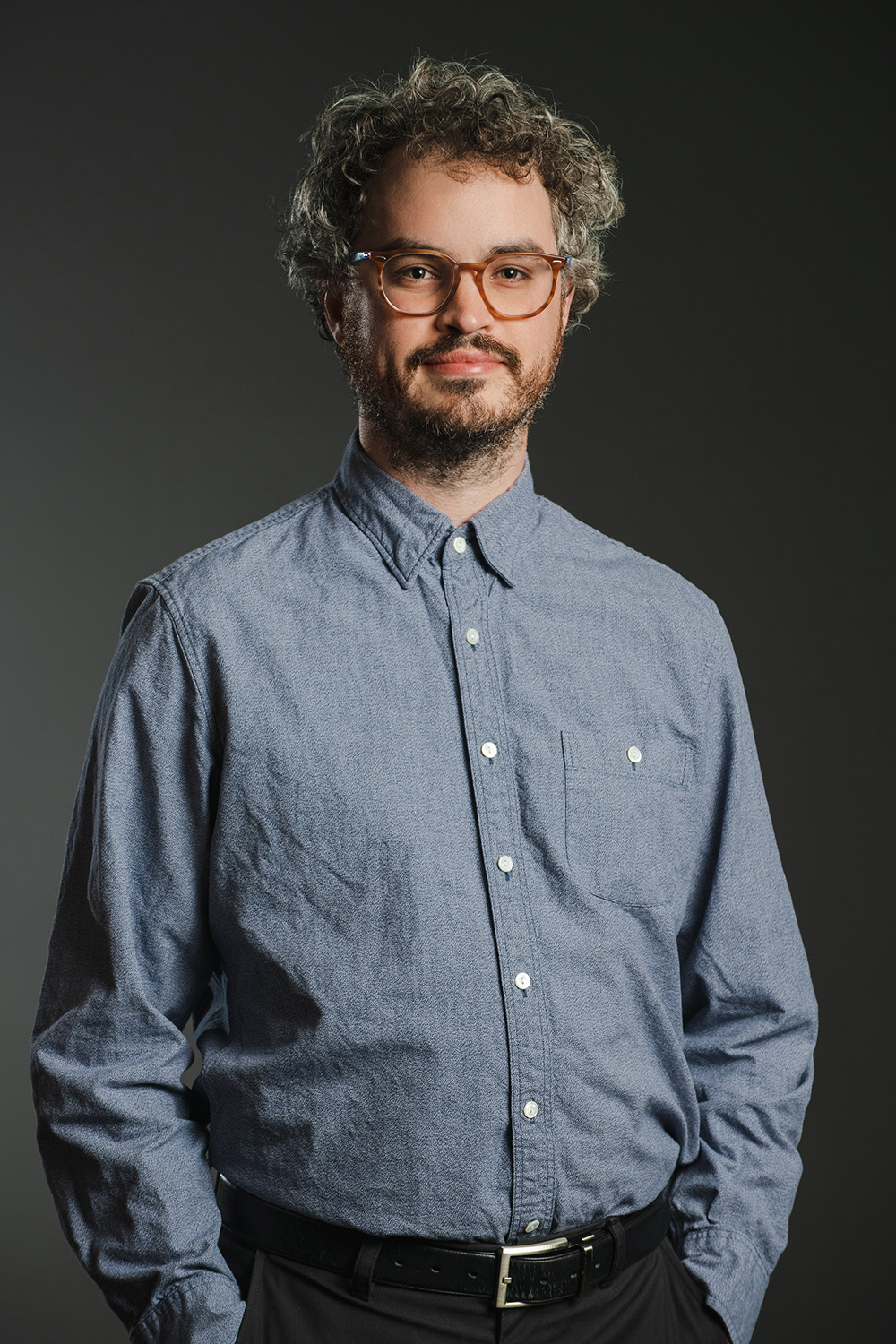 Color photograph of a light skinned man wearing glasses a blue button-up shirt; he looks at the camera with his hands in his pockets