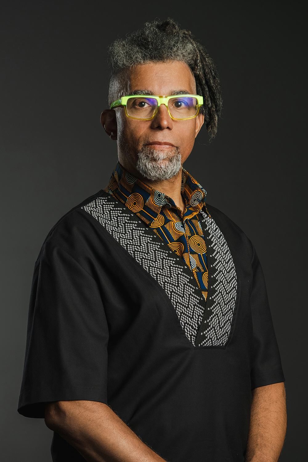 Color photograph of a dark skinned man with gray dreadlocks and goatee wearing glasses and an African inspired shirt, looking directly at the camera