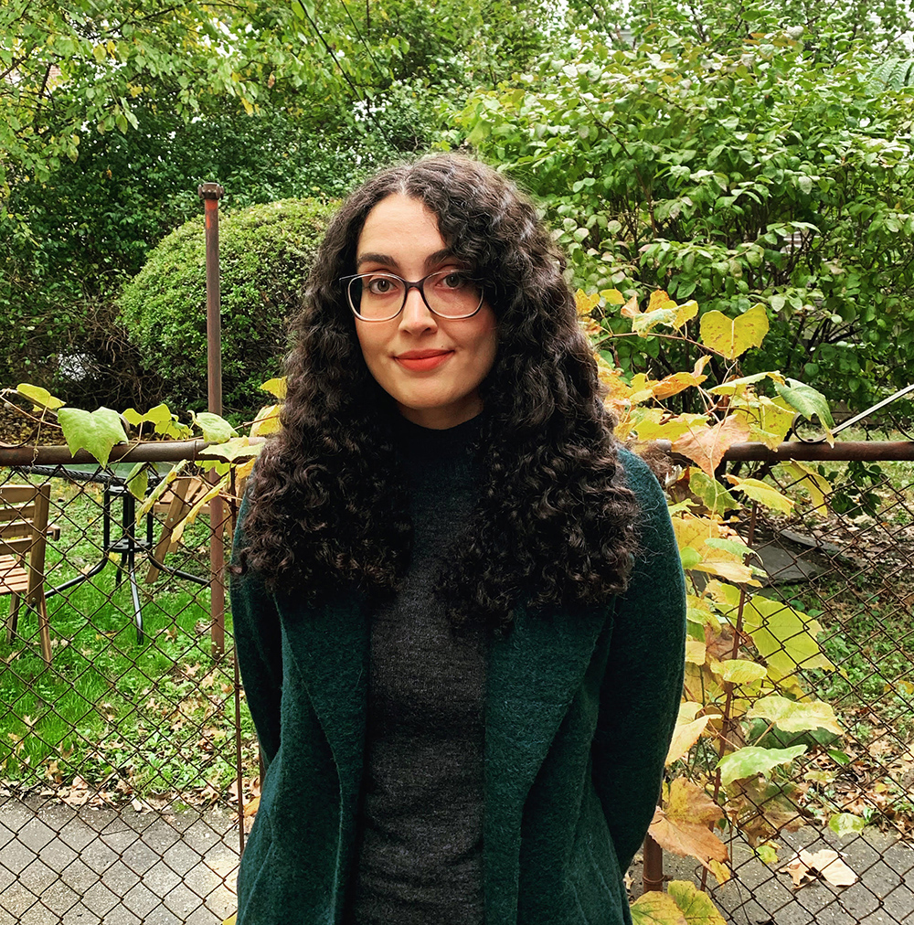 Color photograph of a light skinned woman with dark curly hair wearing glasses and dark clothes; behind her is a chain linked fence, patio furniture, and green shrubs and trees