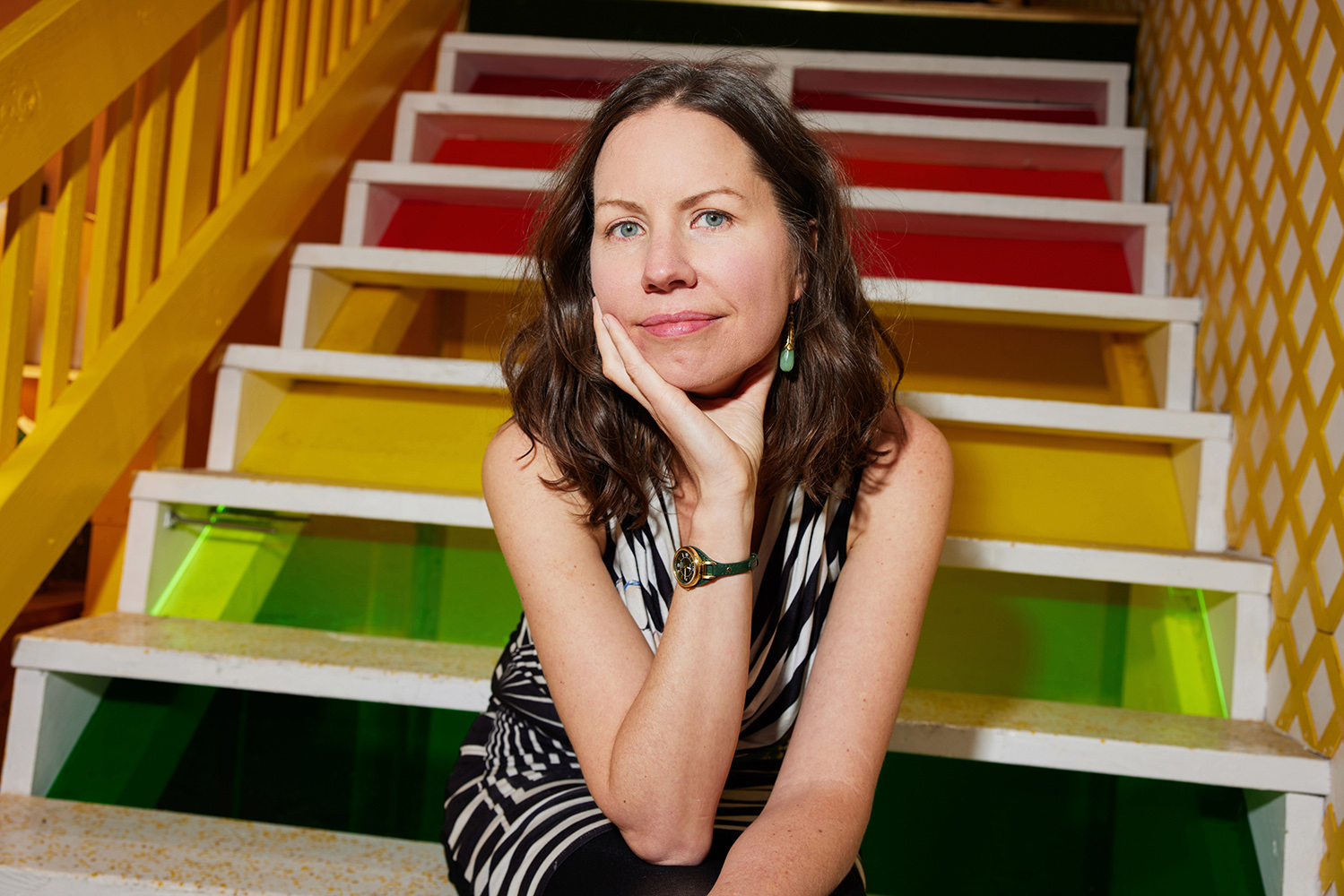 Color photograph of a light skinned woman sitting on a multicolored staircase; she looks at the camera as she rests her head on her hand