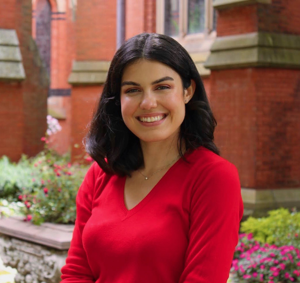 Color photograph of a medium skinned woman with dark hair wearing a bright red sweater; she is outdoors in front of a brick building, smiling at the camera