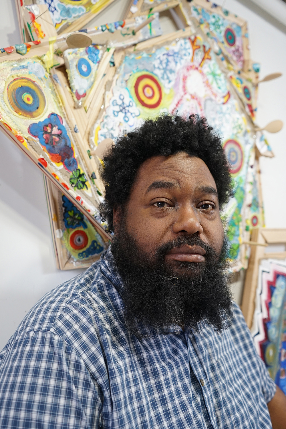 Color photograph of a dark skinned man with a long dark bear and wearing a checkered blue and white shirt; he sits underneath a colorful abstract artwork and looks directly at the camera