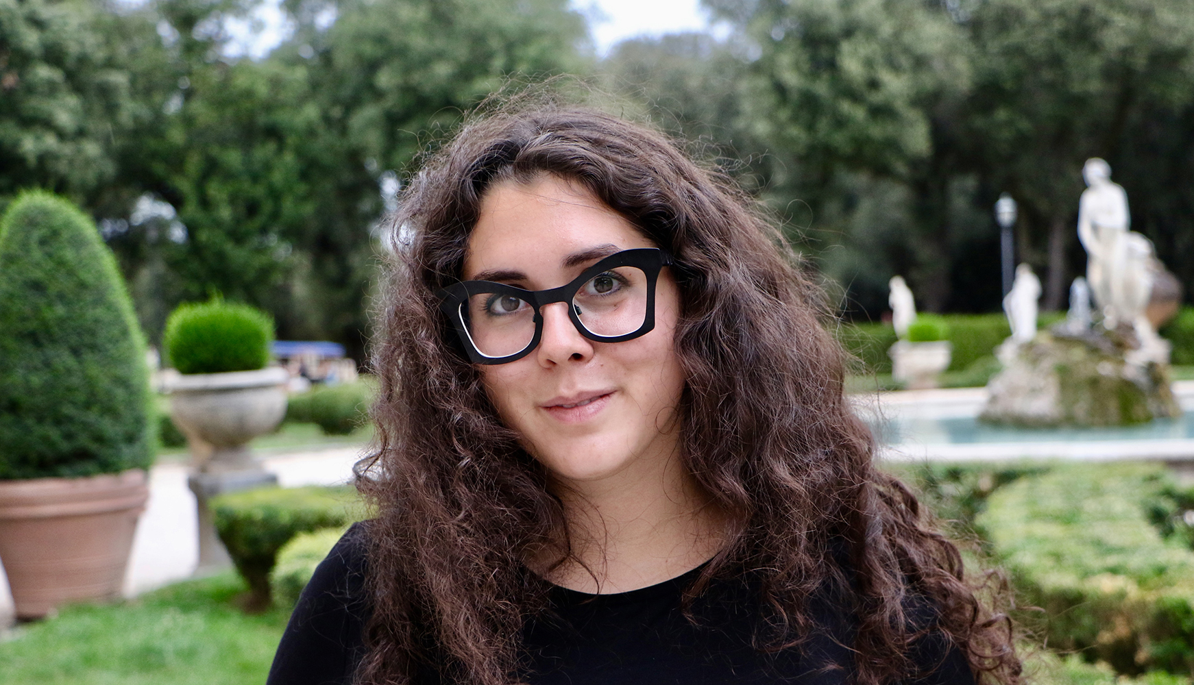 Color photograph of the head of a light skinned woman with brown hair and wearing glasses; green trees can be seen behind her as she looks at the camera