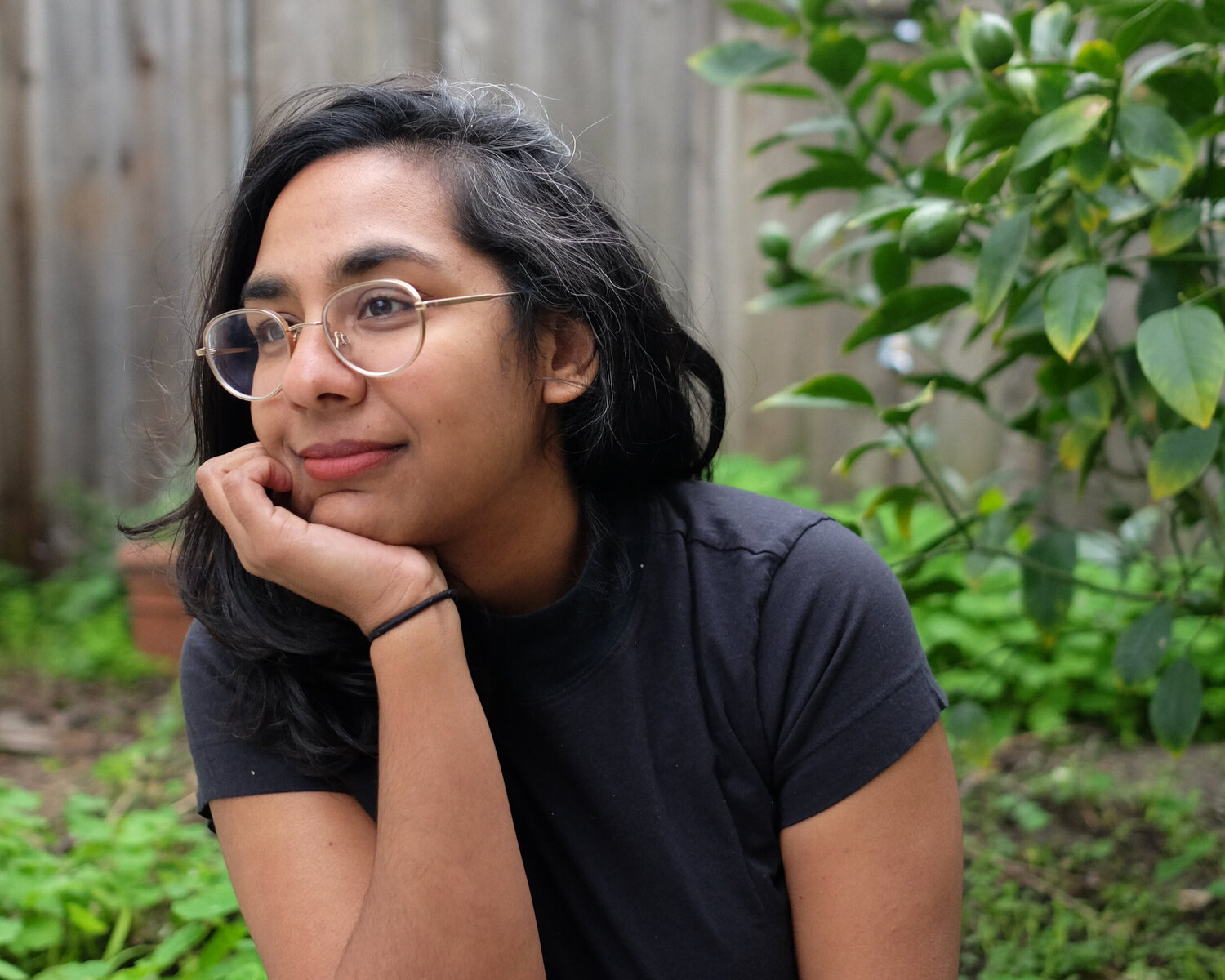 Color photograph of a brown skinned woman with dark hair wearing glasses; she sits in a garden or backyard location with a wooden fence behind her, her hand resting on her chin as she looks off camera